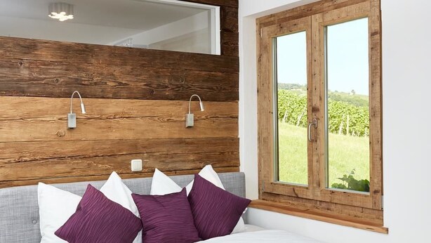 Une chambre à coucher avec une tête de lit en bois, une grande fenêtre donnant sur un vignoble et un lit avec une literie blanche et violette.