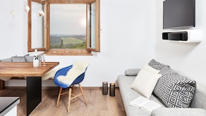 Un salon moderne avec une table à manger en bois, une chaise bleue, un canapé gris, un téléviseur fixé au mur et une fenêtre donnant sur un paysage verdoyant.