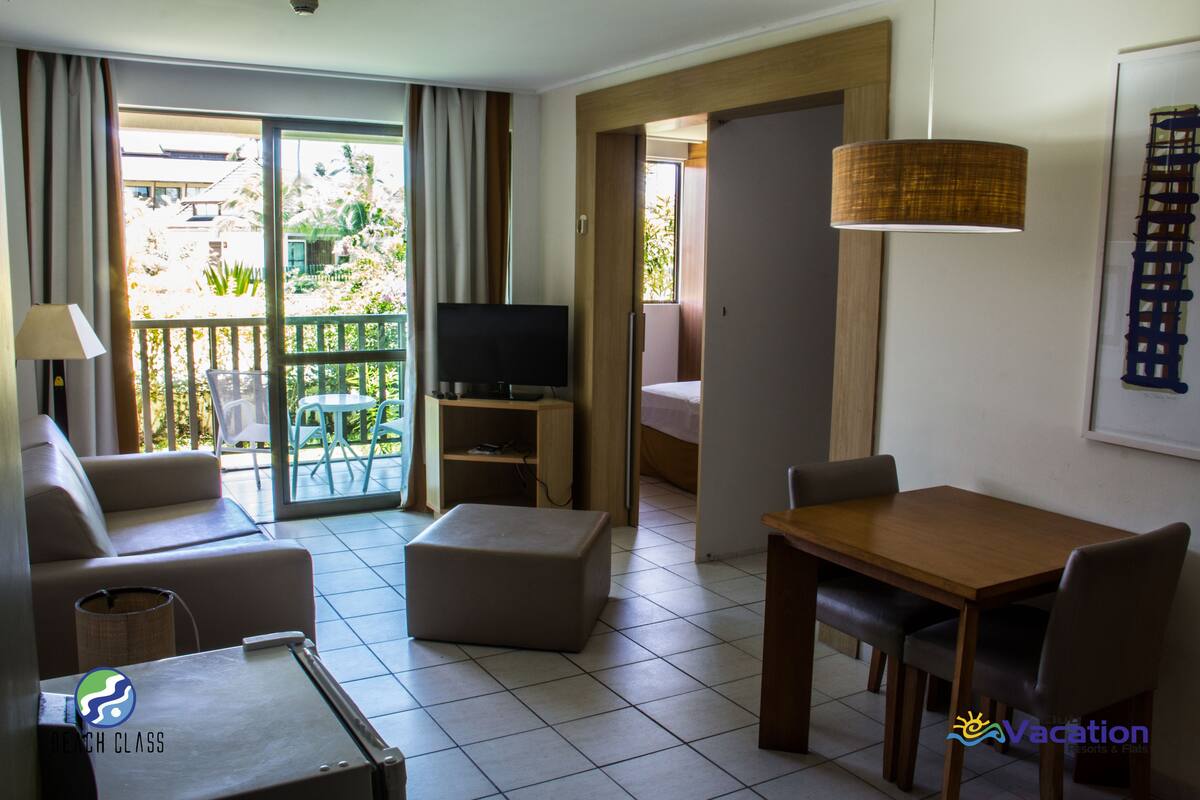 A hotel room with a sofa, a TV, a wooden table, and a balcony with a view of greenery.