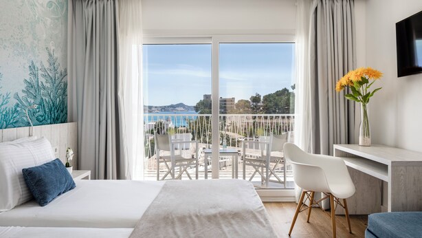 Habitació d'hotel amb llit, escriptori, cadira, televisió i un balcó amb vista al mar i als edificis.