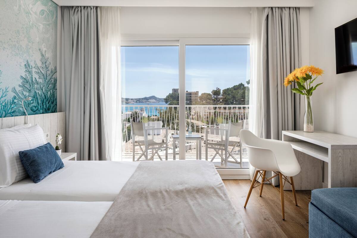 A hotel room with a bed, a desk, a chair, a TV, and a balcony with a view of the sea and buildings.