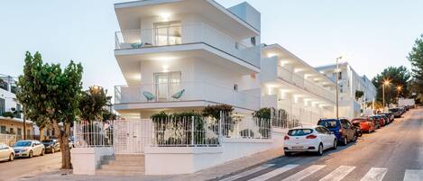 A modern multi-story building with balconies, a pedestrian walkway, and parked cars along the street.