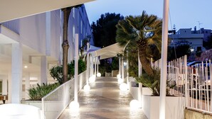 Un passadís cobert amb columnes blanques i llums esfèriques, que condueix a un edifici amb façana blanca i una palmera.