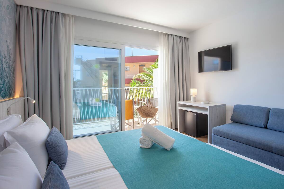 A hotel room with a bed, a desk, a TV, a balcony with a view of buildings and a pool.