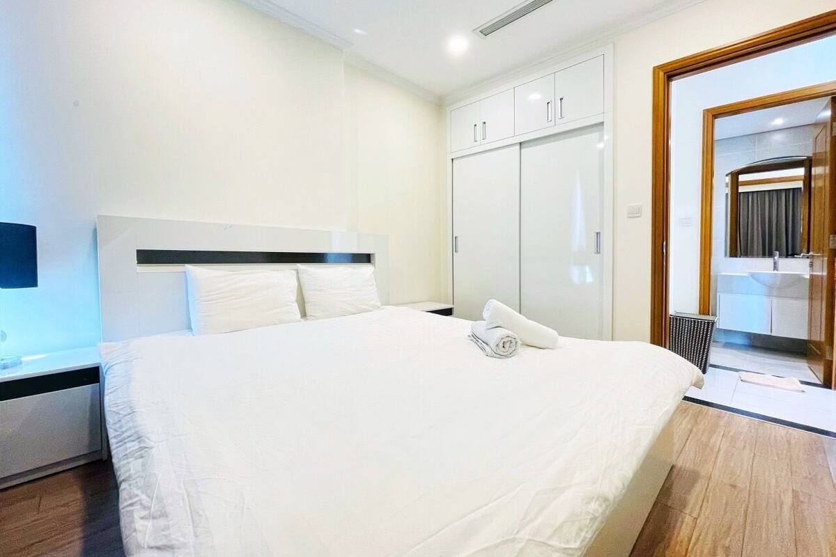 Habitación de hotel con una cama grande, una mesita de noche, un armario y un baño visible a través de una puerta de vidrio.