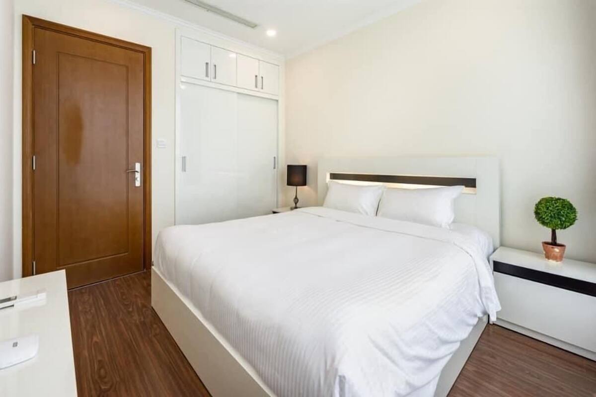Una habitación de hotel con una cama grande, un armario, una mesita de noche con lámpara y una pequeña planta en maceta.