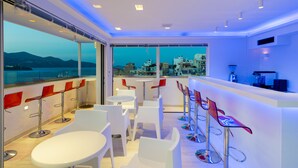Een moderne bar met witte en rode barkrukken, grote ramen met uitzicht op de stad, en een balkon met uitzicht op de bergen.