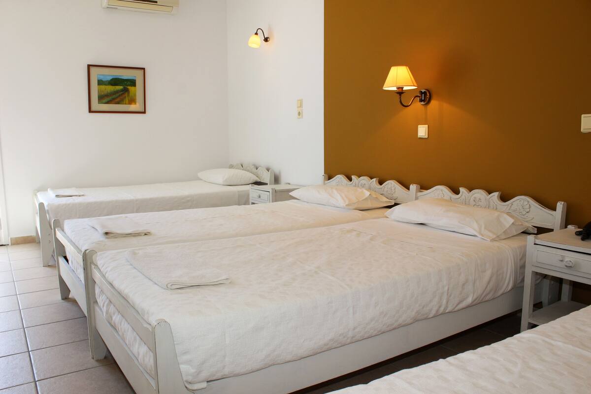Habitación de hotel con tres camas individuales, una mesita de noche con lámpara, una mesita de noche más pequeña con teléfono, y un aplique de pared.
