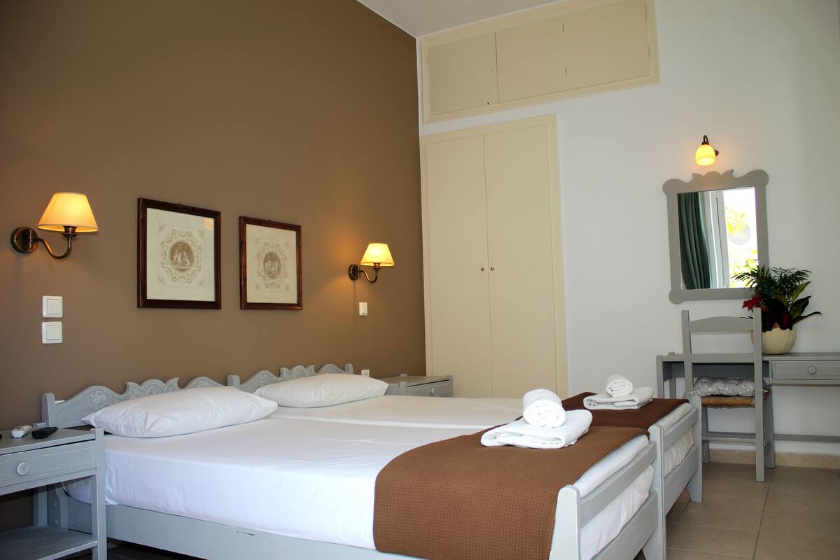 Habitación de hotel con dos camas, un escritorio y una silla. Se observan cuadros enmarcados en la pared, un espejo y una planta en maceta.