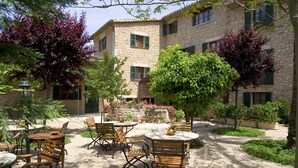 Un patio esterno con tavoli e sedie in legno, circondato da edifici in pietra e rigogliosa vegetazione.