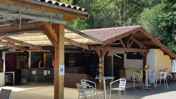 A rustic outdoor café with a wooden pergola, metal chairs, and a snack bar. - Camping Les Vignes (Puy-l'Eveque)