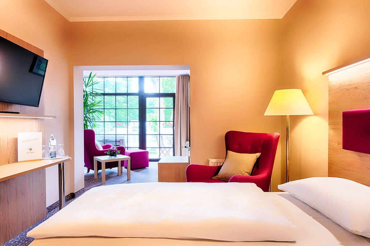 Habitación de hotel con cama, televisor, escritorio con lámpara, una silla roja y ventana con vistas a zonas verdes.