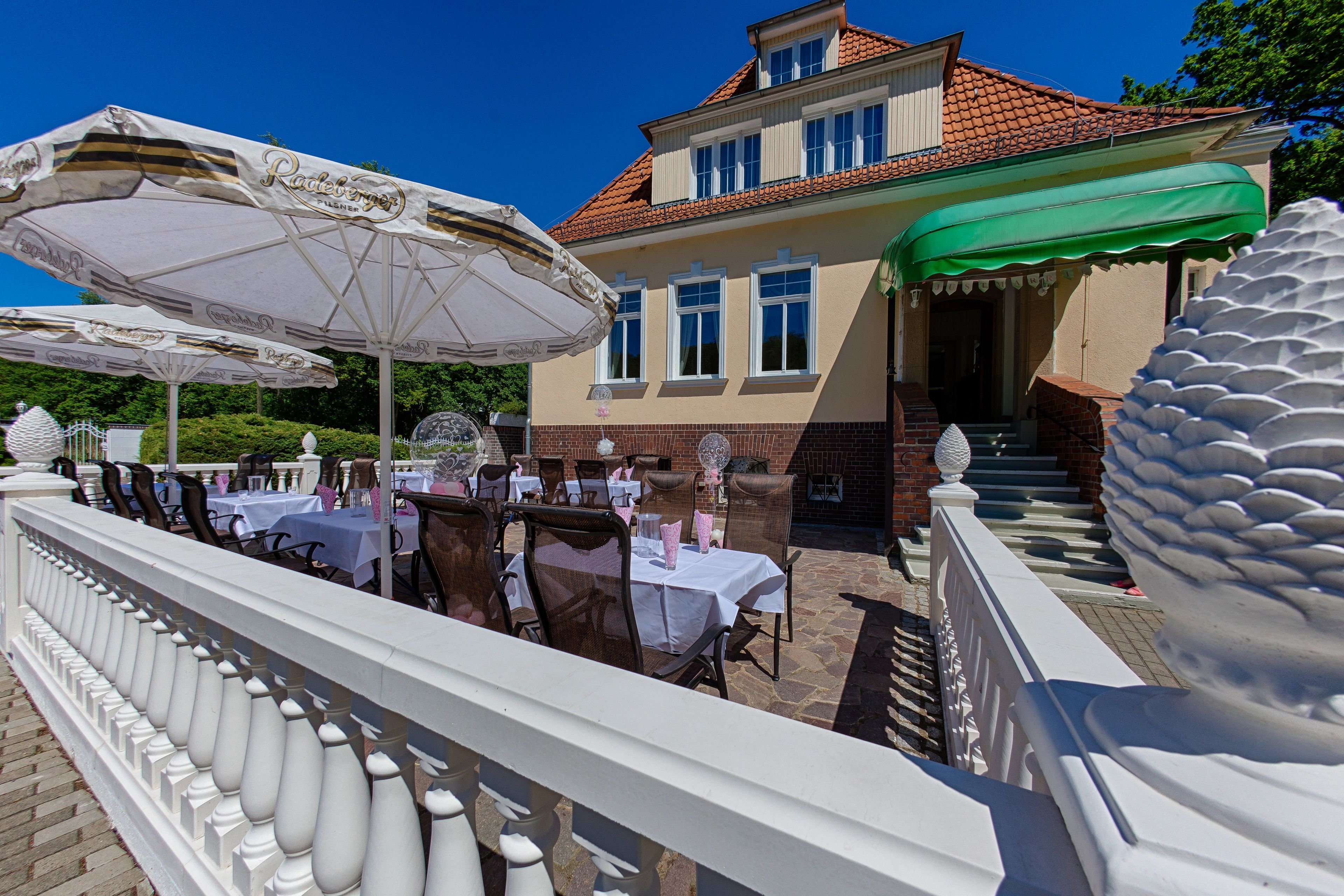 An outdoor dining area with tables and chairs, a white balustrade, and a building with a red-tiled roof.