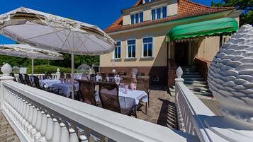 An outdoor dining area with tables and chairs, a white balustrade, and a building with a red-tiled roof.