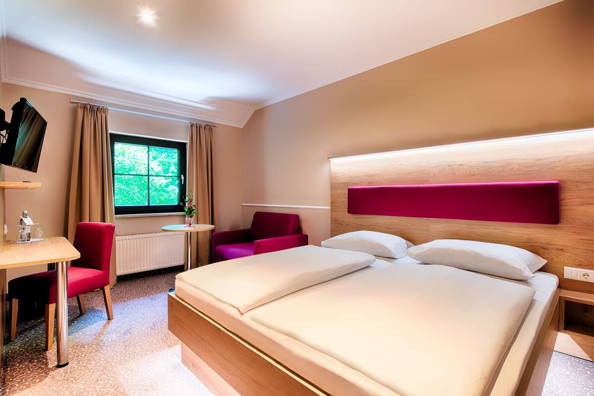 Habitación de hotel con una cama grande, un escritorio con silla, una mesita y un ventanal con cortinas.