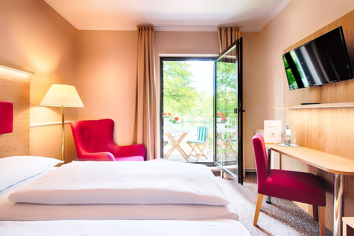 Habitación de hotel con una cama, dos sillas rojas, un escritorio, un televisor de pantalla plana y un ventanal de vidrio que da a un espacio exterior con una mesa y sillas.