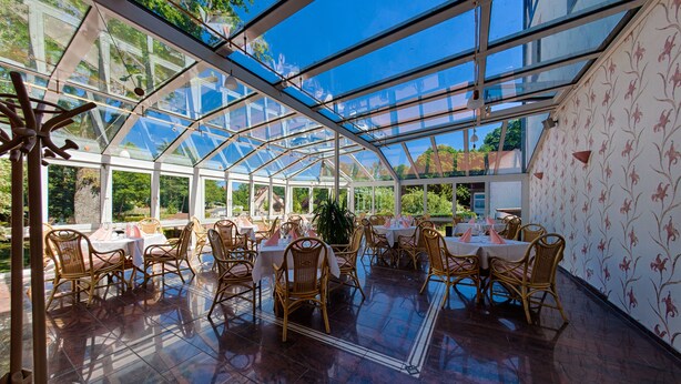 A greenhouse-style dining area with tables and wicker chairs, surrounded by glass walls and a wooden floor.