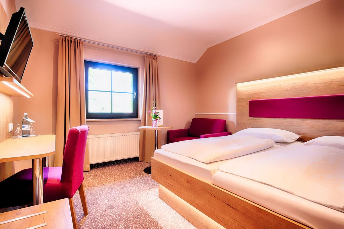 Habitación de hotel con cama, escritorio, silla, televisión y ventana con cortinas.