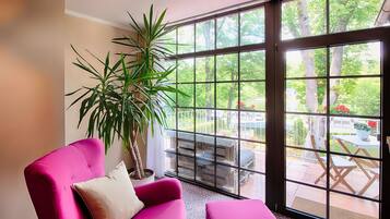 A room with a large window, a pink chair, a potted plant, and a view of an outdoor patio.