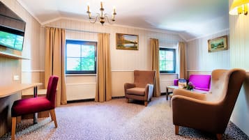 A hotel room with a wooden desk, two armchairs, a sofa, a TV, and a chandelier.