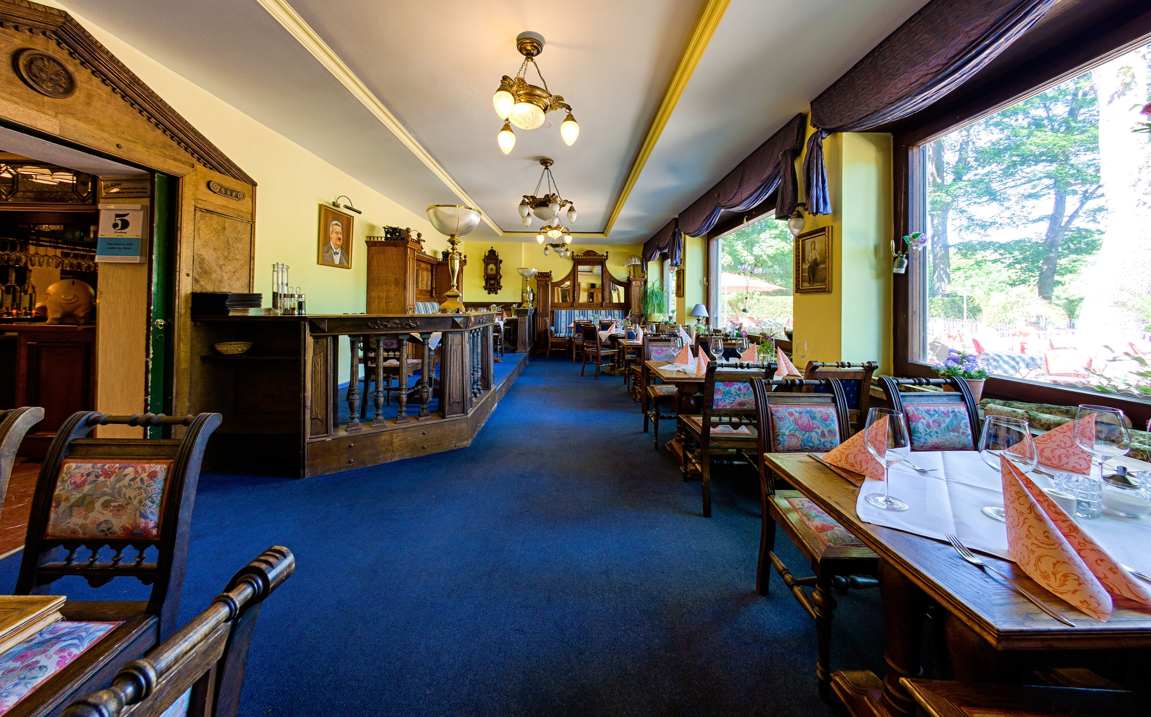A restaurant with a long counter, wooden chairs, and tables. There are hanging lights and a view of trees outside.
