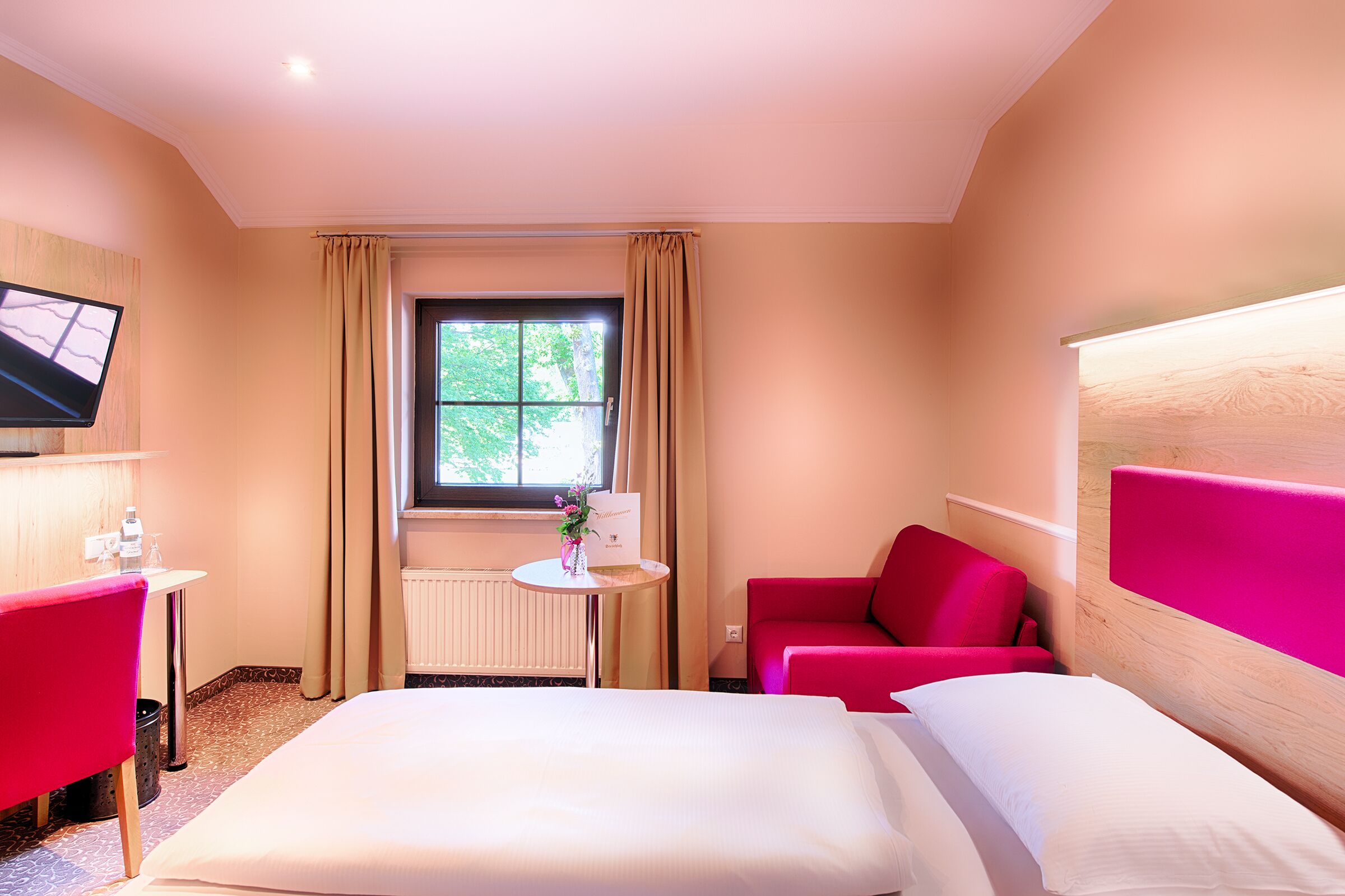 Ein Hotelzimmer mit einem Bett, einem roten Sessel, einem kleinen Tisch mit einer Vase, einem Fernseher an der Wand und einem Fenster mit Vorhängen.