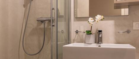 Une salle de bain avec une douche à parois de verre, un lavabo blanc et un miroir au-dessus. On trouve un porte-serviettes au-dessus du lavabo et une petite orchidée en pot posée sur le lavabo.
