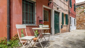 An outdoor seating area with a table and chairs, potted plants, and a building with a red facade and green shutters. - Biennale Garden Art & Architecture Venue (Venice)
