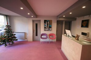 A reception area with a Christmas tree, two chairs, a painting, and a reception desk with a computer and a small decorative item. - Casa vacanze Cucciolo (Rimini)