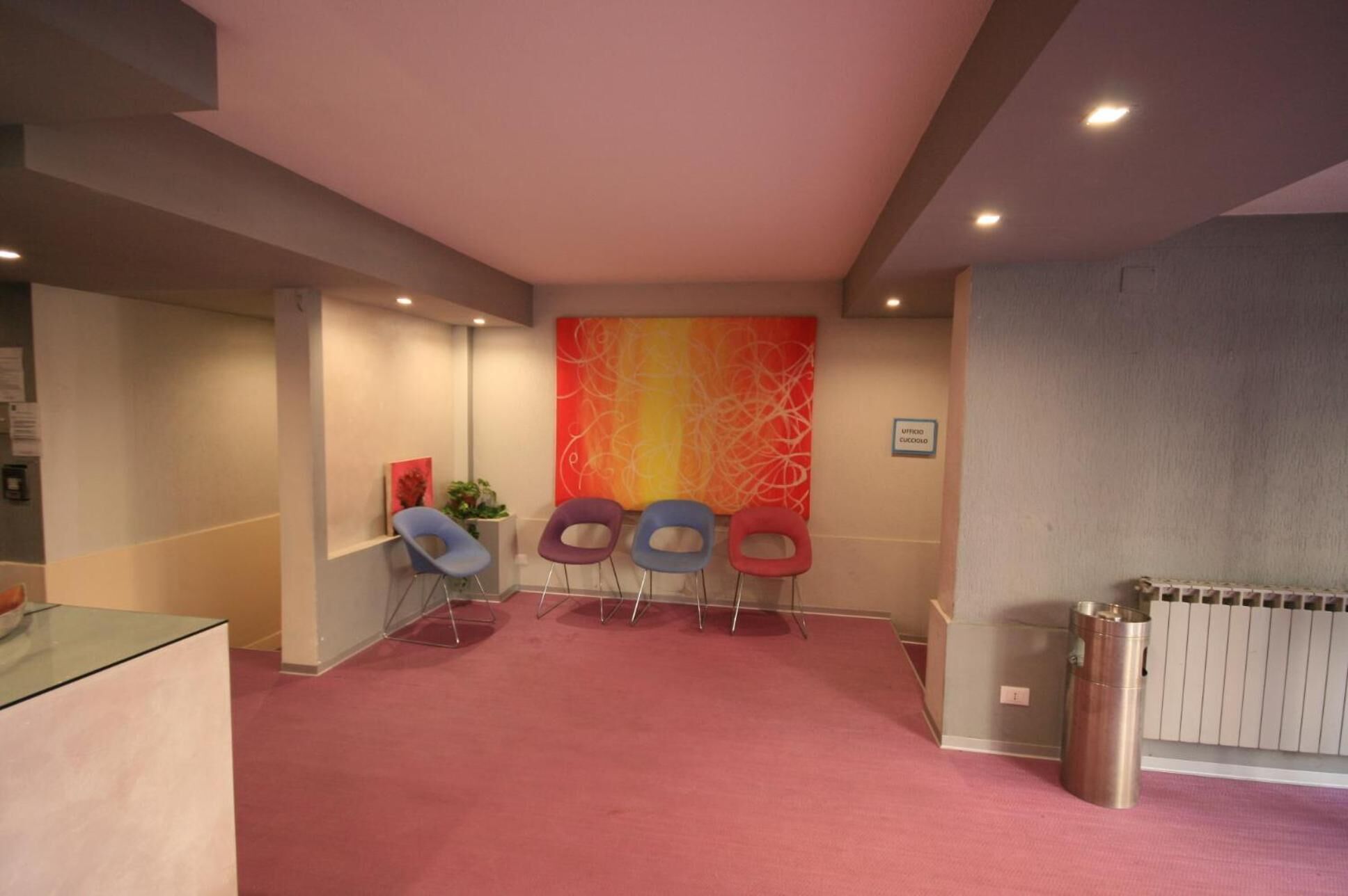 Un espace d’accueil avec un tapis rouge, quatre chaises colorées, un grand tableau abstrait et un radiateur.