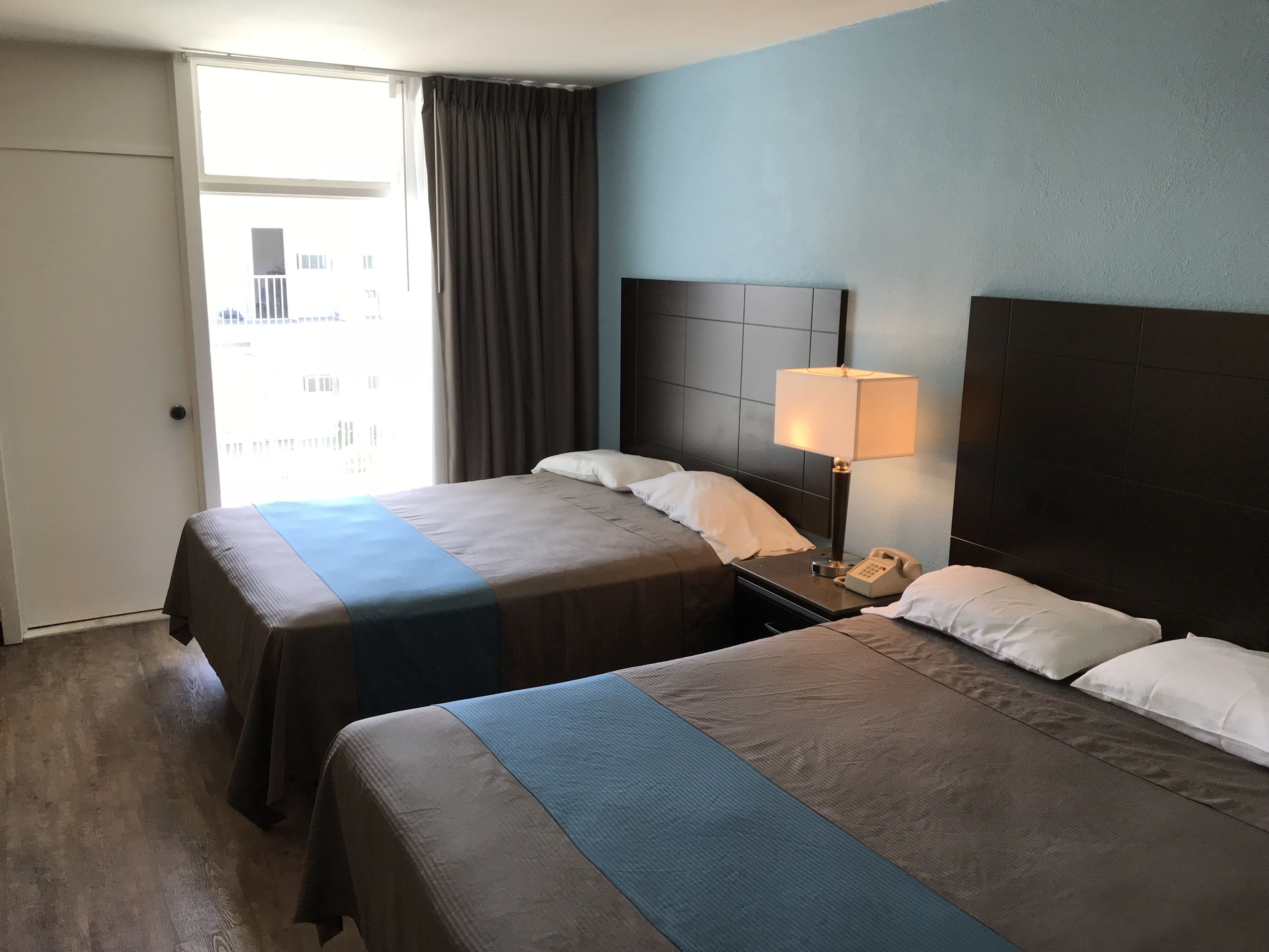 Een hotelkamer met twee bedden, een nachtkastje met een lamp en uitzicht op gebouwen door een raam.