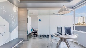 A modern office space with a glass partition, two desks with computers, a bench, and a hanging light fixture. - One Puebla Angelópolis Periférico (San Andrés Cholula)