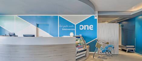 Un área de recepción con un mostrador curvo, una pared con el logo 'one', una bicicleta y una zona de estar.