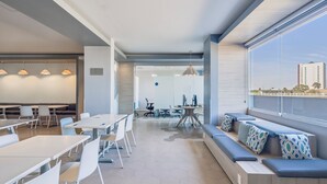 A modern hotel lobby with a dining area, seating by the window, and a visible office space in the background. - One Puebla Angelópolis Periférico (San Andrés Cholula)