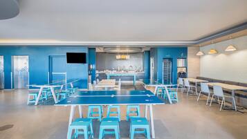 Un comedor moderno con mesas y sillas azules y blancas, una cocina al fondo e iluminación colgante.