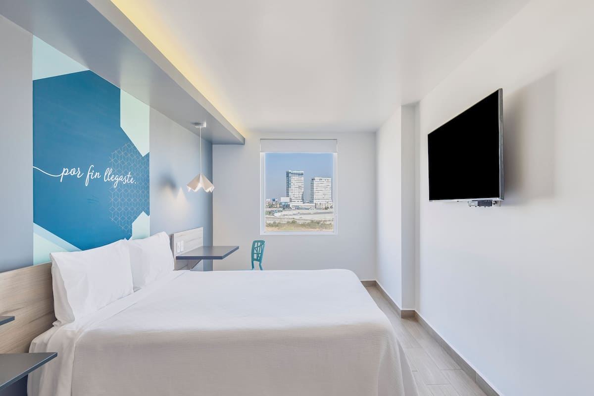 Una habitación de hotel moderna con una cama, un escritorio, una silla, un televisor y un ventanal con vista a la ciudad.