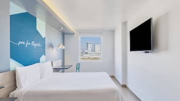 A modern hotel room with a bed, a desk, a chair, a TV, and a window with a city view.