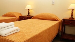 A hotel room with two single beds, each with a matching pillow and a wooden nightstand with a lamp. A stack of white towels is placed on the bed. - Hotel Universal (Mendoza)