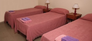 Ein Zimmer mit drei Einzelbetten, jedes mit einer rosa Tagesdecke und einem violetten Handtuch. Zwei Nachttische mit Lampen sind vorhanden.