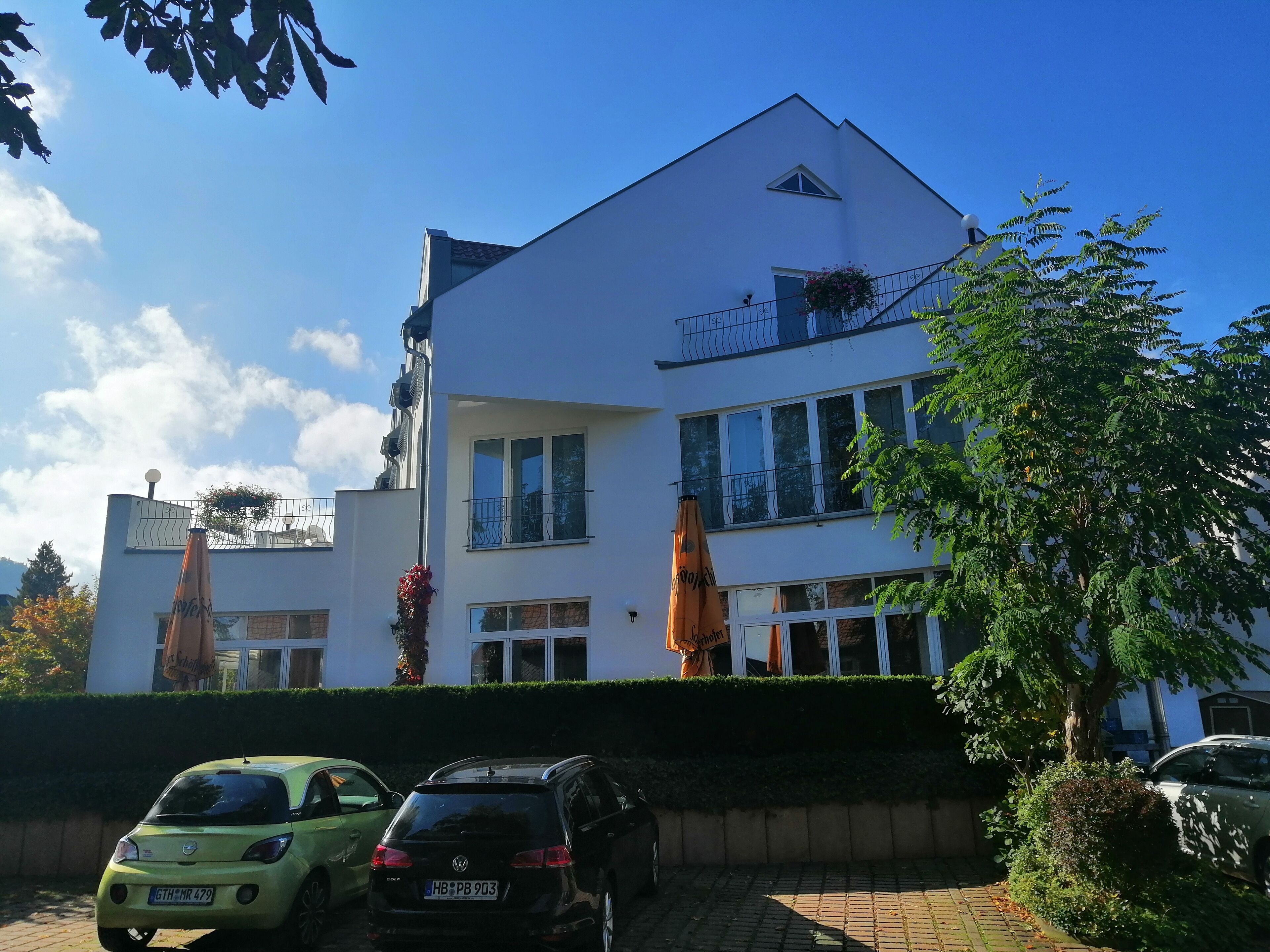 A two-story building with a white facade, multiple windows, and a balcony with a railing. There are two cars parked in front, and the building is surrounded by greenery.