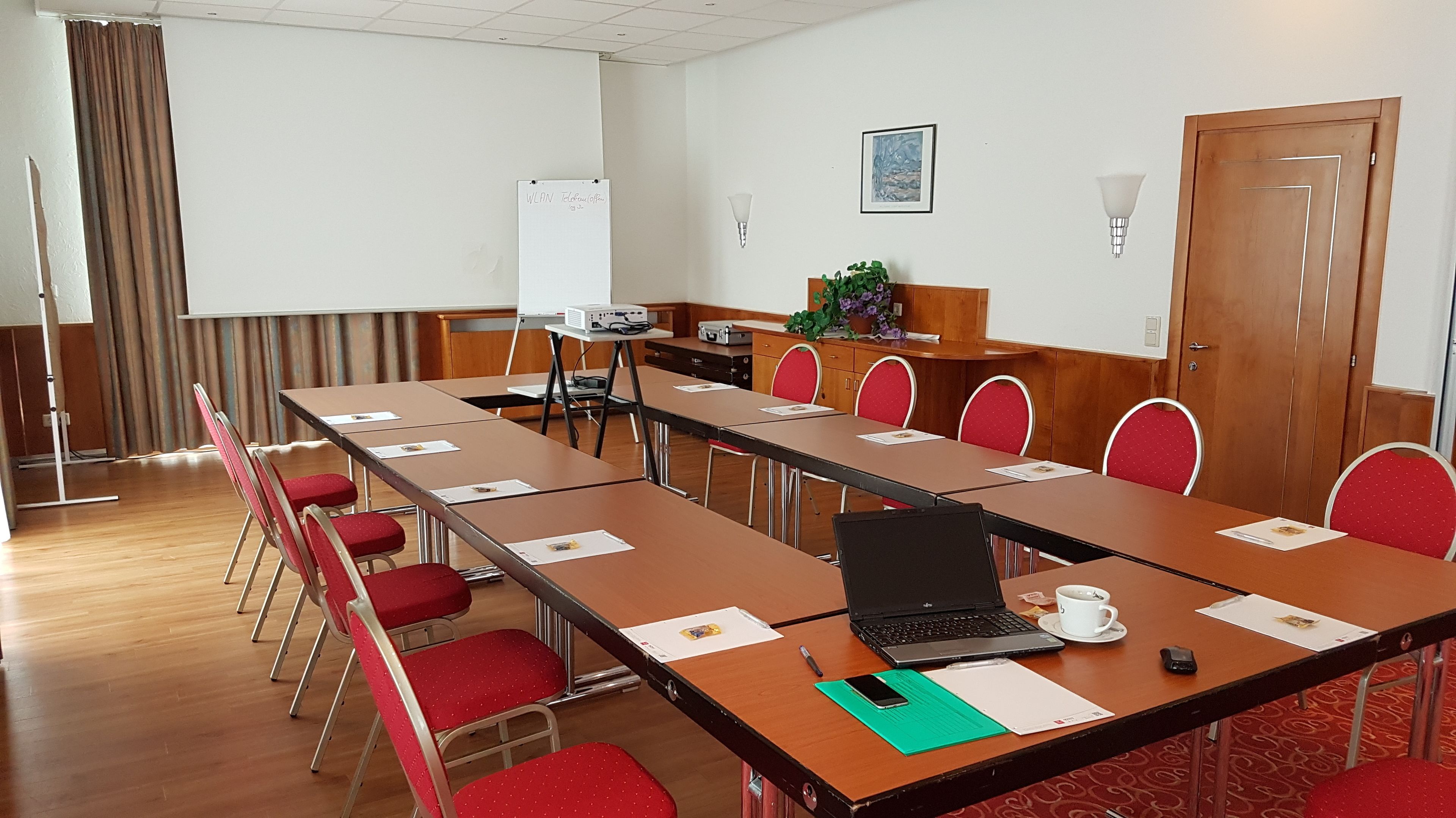 A conference room with a long table, red chairs, a laptop, a projector, and a whiteboard.