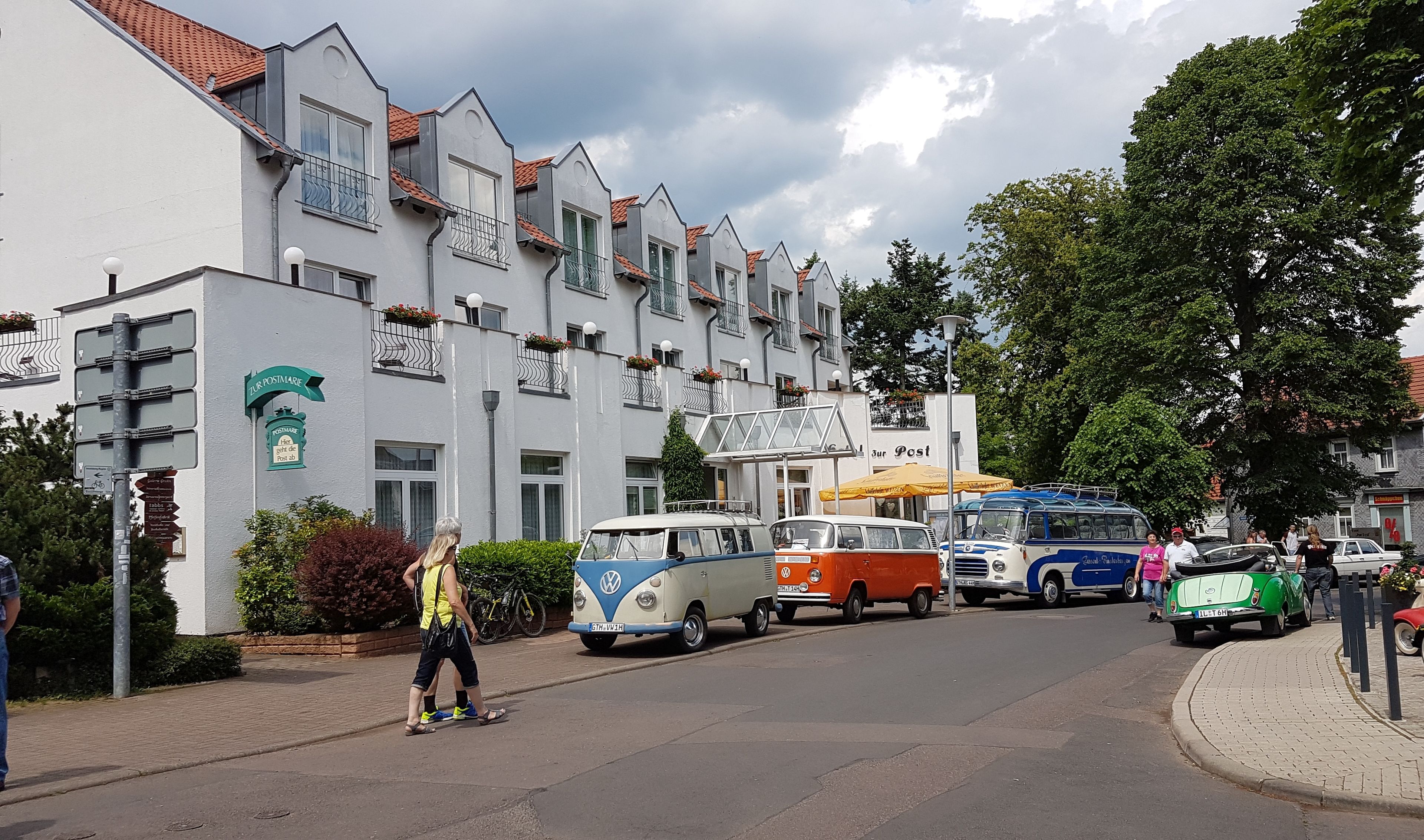 A street with vintage cars parked along the side, a building with a sign reading "Zur Post", and a pedestrian walking by.