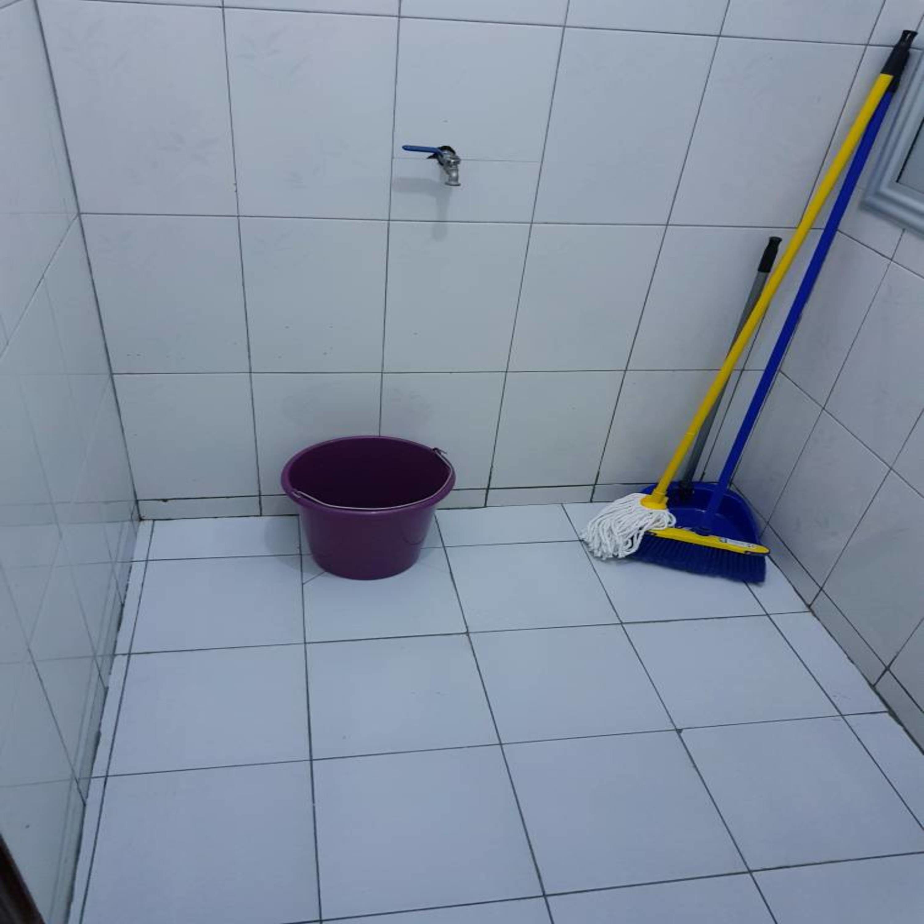 A small, tiled bathroom with a purple bucket, a mop, and a showerhead.