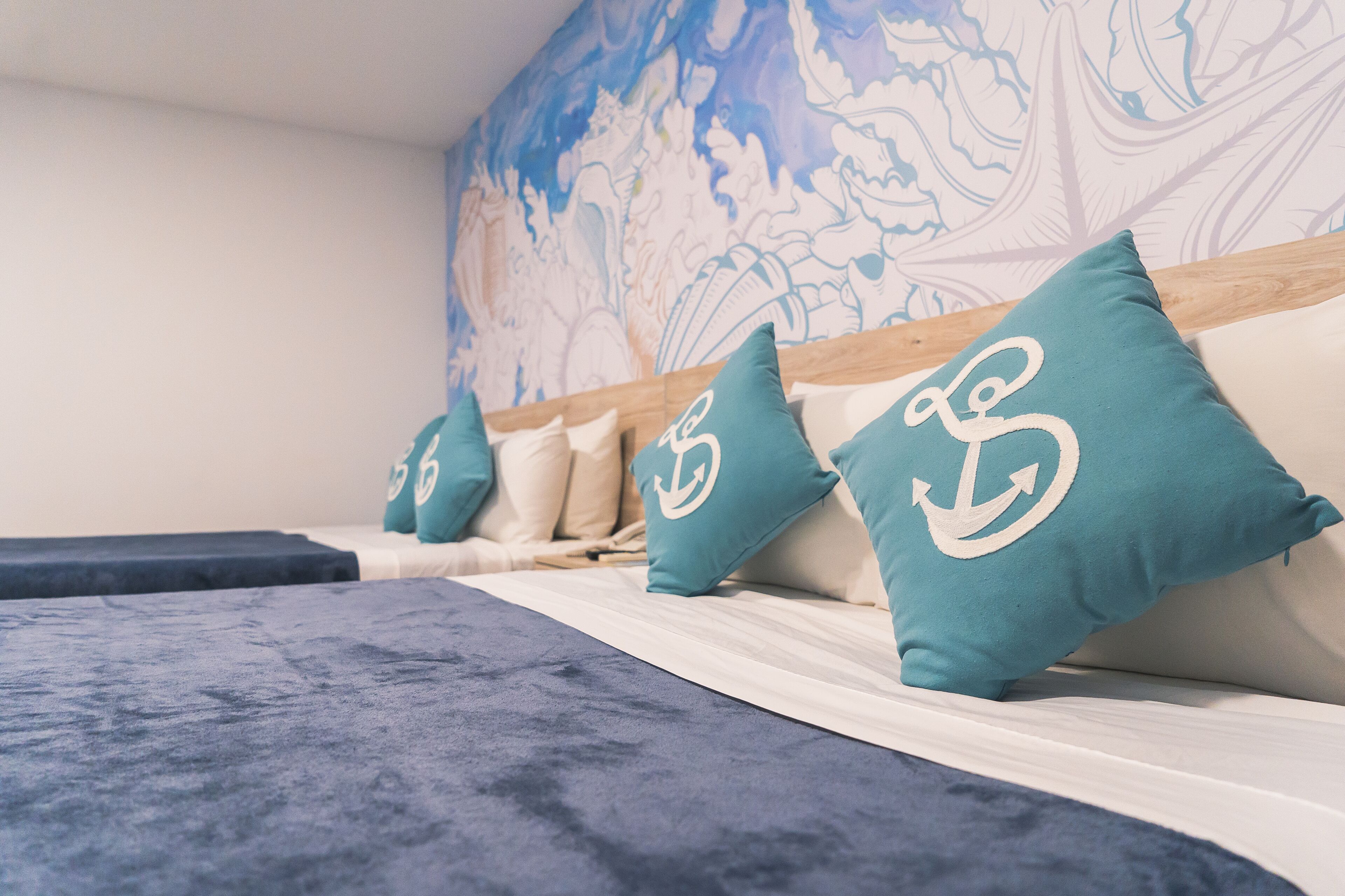 A hotel room with a bed, teal pillows with anchor designs, and a mural on the wall.