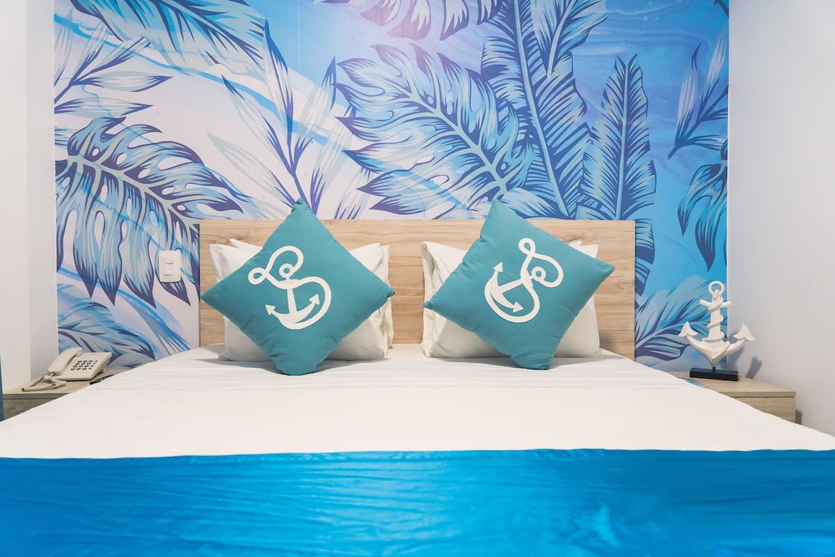 A bed with two teal pillows featuring anchor designs, a wooden headboard, a blue bedspread, and a wall mural of blue and white leaves.