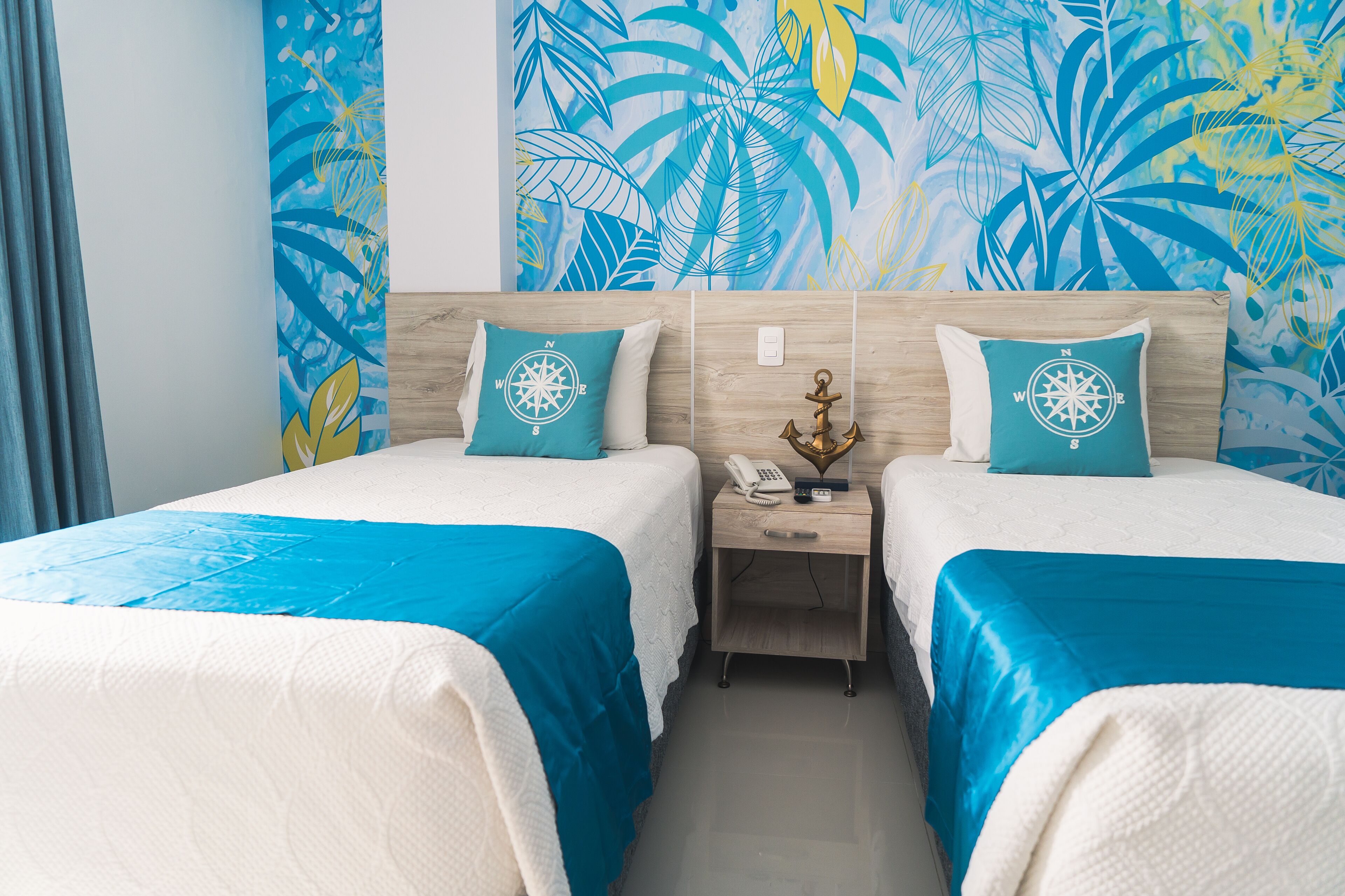 A hotel room with two beds, each with a blue bedspread and a white pillow with a compass design. There is a wooden nightstand between the beds with a phone and a decorative item.