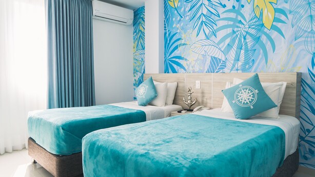 A hotel room with two beds, a large window with curtains, an air conditioning unit, and a tropical leaf-patterned wall. - Sea Colors Hotel (San Andrés)