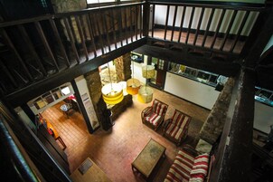 A spacious loft-style living area with a high ceiling, exposed wooden beams, and a stone fireplace. There are multiple seating areas with patterned cushions and a central coffee table.