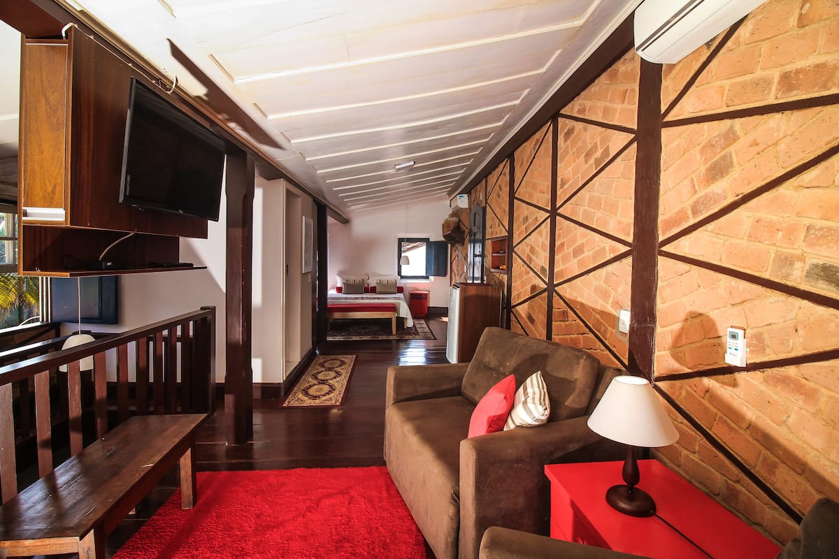 Uma sala de estar acolhedora, com um banco de madeira, um tapete vermelho e um sofá marrom com abajur. Uma televisão está fixada na parede, e visível está uma parede de tijolos com um padrão geométrico.