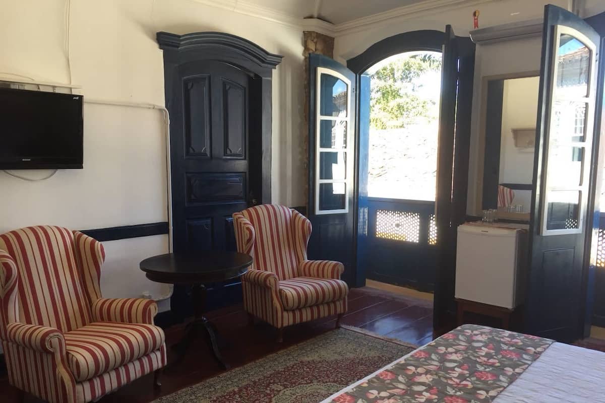 Um quarto com uma cama, duas poltronas listradas, um armário de madeira escura e uma pequena mesa.